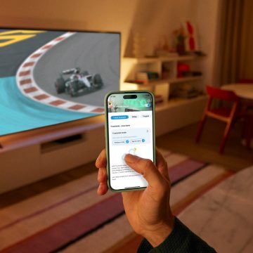 Eine Hand, die ein Smartphone mit der WiZ App in der Hand hält. Ein Fernseher zeigt ein Rennen; der TV-Hintergrund wird beleuchtet mit der smarten WiZ Beleuchtung.