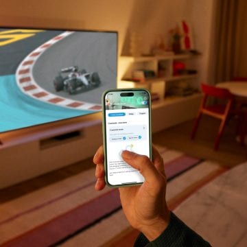 Eine Hand, die ein Smartphone mit der WiZ App in der Hand hält. Ein Fernseher zeigt ein Rennen; der TV-Hintergrund wird beleuchtet mit der smarten WiZ Beleuchtung.