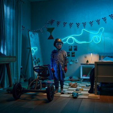 Ein junger Junge steht neben einem Go-Kart in seinem Zimmer, das von einem blauen WiZ Smart Flex Strip in Form eines Rennwagens beleuchtet wird.