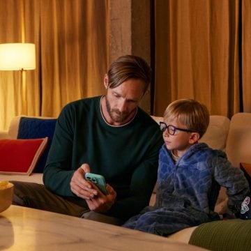 Un padre mostrando su teléfono a su hijo pequeño en el sofá. Una lámpara con una bombilla inteligente WiZ proyecta una luz cálida.